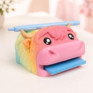 May include: A rainbow-colored highland cow desk organizer featuring a pink face, tan horns, and a textured coat. The figure has a slot for a blue pen across the top and a horizontal opening at the mouth that holds a stack of blue sticky notes.