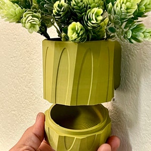 Könnte beinhalten: Ein grüner Keramiktopf mit strukturierter Oberfläche. Der Topf ist in zwei Teile geteilt, wobei der obere Teil abnehmbar ist. Der Topf ist mit einer grünen Kunstpflanze gefüllt.