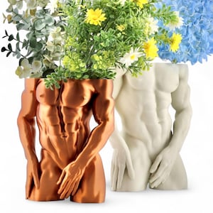 May include: Two decorative planters shaped like muscular male torsos. One is bronze-colored, the other is white. Both planters are filled with colorful artificial flowers and greenery, including yellow, white, and blue blooms. The planters are set against a white background.