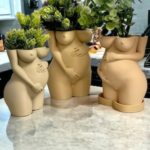 May include: Three nude-colored ceramic planters shaped like pregnant figures. Each planter has a different pose and is holding their belly. The planters are filled with green plants.