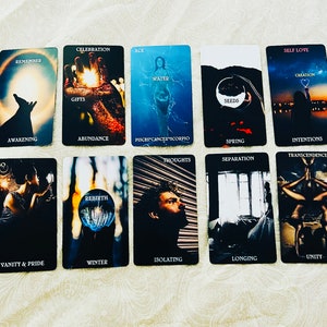Op de afbeelding: Een verzameling tarotkaarten met diverse illustraties en tekst. De kaarten tonen afbeeldingen van handen, figuren en natuurlijke elementen, met woorden als "Remember", "Celebration" en "Self Love". Het kleurenpalet is donker met accenten van blauw en goud.