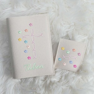 May include: Beige Bible cover and matching pouch, both embroidered with a pink fish cross and colorful flowers. The cover has the name "Tabea" in green script. The items are on a white, fluffy surface.