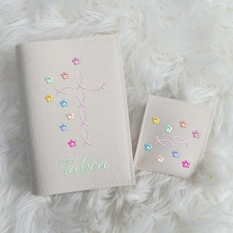 May include: Beige Bible cover and matching pouch, both embroidered with a pink fish cross and colorful flowers. The cover has the name "Tabea" in green script. The items are on a white, fluffy surface.