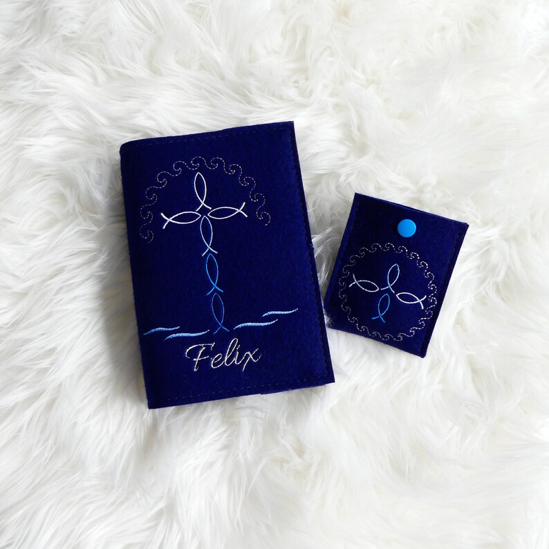 May include: Navy blue felt book cover and small pouch set. Both items feature an embroidered cross design made of fish, with the name "Felix" embroidered on the book cover. The pouch has a blue button closure.