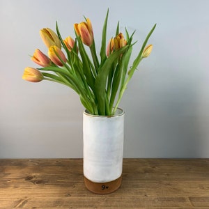 May include: A white ceramic vase with a brown base, holding a bouquet of yellow and orange tulips. The vase has a handwritten inscription that reads "9".