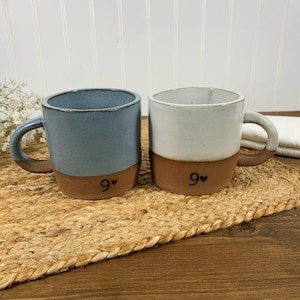 May include: Two ceramic mugs with brown bottoms and a white or blue glaze. The mugs have a black heart and the number 9 on the bottom. The mugs are sitting on a woven brown mat.