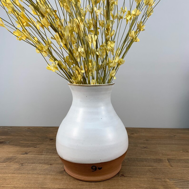 May include: White ceramic vase with a brown base. The vase has a rounded body and a narrow opening. It is filled with dried yellow flowers. The base of the vase is marked with the number "9" and a heart.