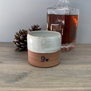 May include: A white and brown ceramic mug with the number "9" and a heart symbol on the bottom half. The mug is sitting on a wooden surface.