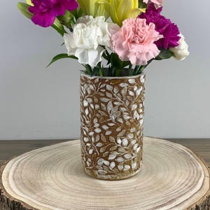 May include: A brown vase with a leaf pattern holds a bouquet of flowers. The flowers include pink, white, and purple carnations, and yellow lilies. The vase is cylindrical and sits on a wooden slice.