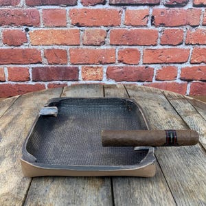 May include: A square, textured ashtray with a cigar resting on its edge. The ashtray is dark brown with a lighter brown rim. The cigar is brown with a black and red band. The background is a brick wall.