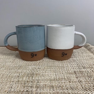 May include: Two ceramic mugs. One is light blue, the other is white. Both have a brown base and handle. The number "9" with a heart is printed on the base of each mug. The mugs are on a woven surface.