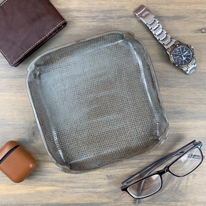 May include: A square, gray ceramic tray with a textured surface. A brown leather wallet, a silver wristwatch, a brown AirPod case, and black-framed glasses are arranged around the tray on a wooden surface.