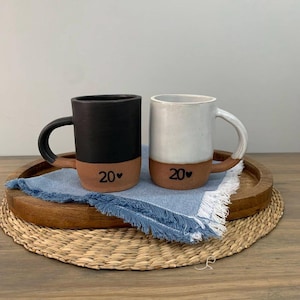 May include: Two ceramic mugs, one black and one white, with brown bases and handles. Each mug has the number "20" with a heart symbol. The mugs sit on a wooden tray with a blue fringed cloth.