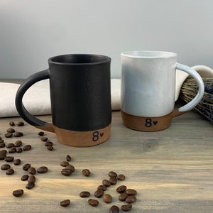 May include: Two ceramic mugs with brown rims and a black heart with the number 8 on the bottom. One mug is black and the other is white. The mugs are on a wooden surface with coffee beans scattered around them.