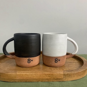 May include: Two ceramic mugs with handles, one featuring a matte black upper section and the other a white upper section. Both mugs have a natural terracotta-colored base and are marked with the black text 8 followed by a small heart symbol.