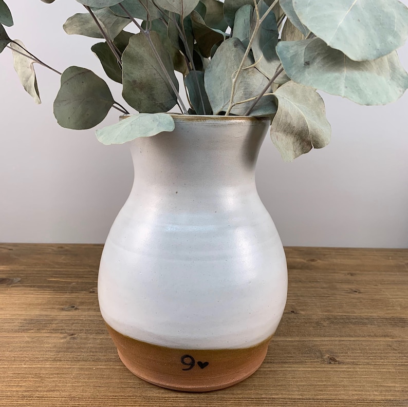 May include: White ceramic vase with a brown rim and a brown base. The vase has a rounded body and a narrow neck. The base is marked with the number "9" and a heart.