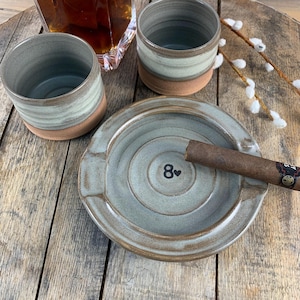 May include: A handmade ceramic ashtray with a cigar, two matching tumblers, and a decanter of amber liquid. The ashtray is light blue-green with the number 8 and a heart symbol. The tumblers have a light blue-green top and a brown base.