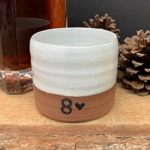 May include: A handmade ceramic tumbler with a white upper half and a reddish-brown base. The number "8" and a heart symbol are printed on the base. The tumbler is cylindrical and appears to be suitable for beverages.