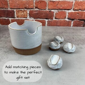 May include: A ceramic ashtray and matching small round pieces. The ashtray is light gray with a brown base and a curved cutout. The small pieces are white with a brown base. Text on the image says "Add matching pieces to make the perfect gift set."
