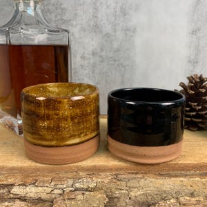 May include: Two ceramic drinking cups, one with a brown and yellow speckled glaze and the other with a black glaze. Both cups have a brown rim.
