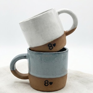 May include: Two stacked ceramic mugs with exposed terracotta bases and glazed upper sections. The top mug is white, and the bottom mug is light blue. Both feature the number 8 followed by a small heart symbol printed in black on the clay base.