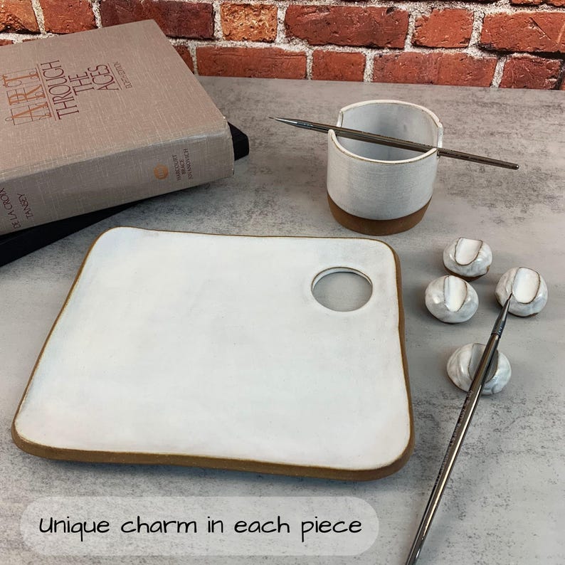 May include: A white ceramic artist's palette with a brown rim and a circular hole, accompanied by a small white cup, paint holders, and paintbrushes. The text "Unique charm in each piece" is at the bottom.