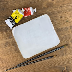 May include: A white ceramic artist's palette with a brown rim, two paintbrushes, and two tubes of paint. One tube is yellow with black text, and the other is red. The palette and brushes are on a wooden surface.