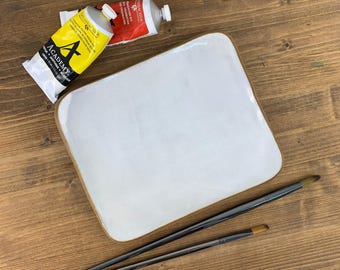 May include: A white ceramic artist's palette with a brown rim, two paintbrushes, and two tubes of paint. One tube is yellow with black text, and the other is red. The palette and brushes are on a wooden surface.