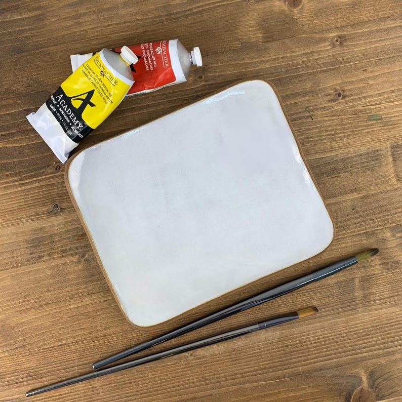 May include: A white ceramic artist's palette with a brown rim, two paintbrushes, and two tubes of paint. One tube is yellow with black text, and the other is red. The palette and brushes are on a wooden surface.
