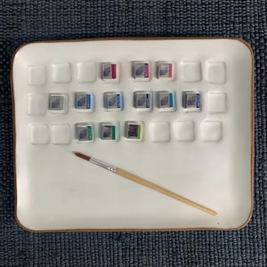 May include: A white ceramic palette tray with multiple rectangular indentations, some containing small watercolor paint pans in various colors. A wooden paintbrush with a brown tip rests diagonally across the center of the tray.