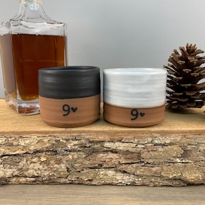 May include: Two ceramic shot glasses with a brown base, one with a black top and one with a white top. Both glasses have the number "9" with a heart symbol written on the bottom.
