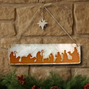 May include: A rectangular Christmas sign with a nativity scene silhouette in orange against a white and gold background. A star hangs above the sign, suspended by twine. The sign is displayed against a brick wall, with a garland of greenery and berries below.
