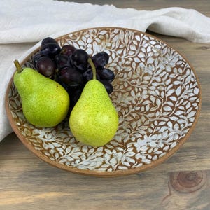 May include: A round, brown ceramic serving bowl with a white leaf pattern. The bowl is filled with green pears and dark purple grapes. The bowl has a raised edge and sits on a wooden surface.