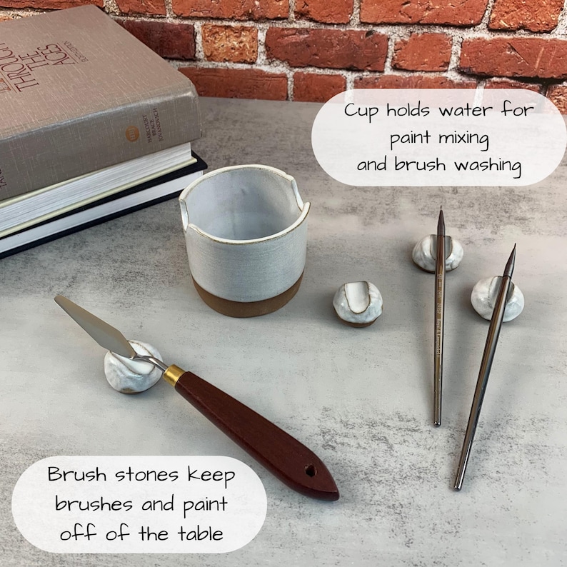 May include: A ceramic paint cup, brush stones, and paint brushes are arranged on a table. The cup holds water for paint mixing and brush washing. Brush stones keep brushes and paint off the table.