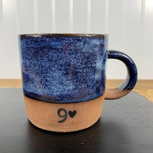 May include: A handmade ceramic mug with a blue speckled glaze on the upper half and a natural clay base. The mug has a curved handle and the number "9" with a heart symbol is painted on the base. The mug is on a black surface.