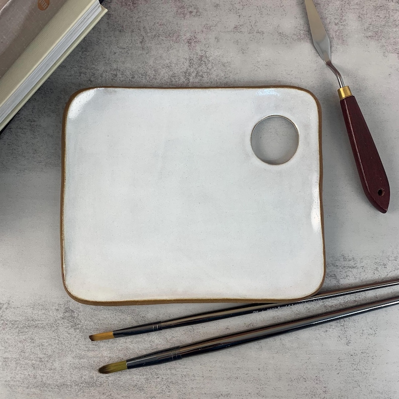 May include: A white ceramic artist's palette with a brown rim and a circular thumb hole. A palette knife with a maroon handle and two paint brushes are also present. The art supplies are arranged on a textured surface.