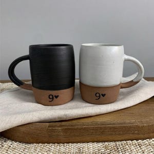 May include: Two ceramic mugs with a two-tone design. One mug features a matte black upper section, while the other has a white upper section. Both mugs have a natural terracotta base with the number 9 and a small heart symbol printed in black.