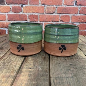 May include: Two green and brown ceramic mugs with a shamrock design. The mugs are handmade and have a rustic look.
