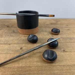 May include: A set of black ceramic paintbrush holders with a brown rim. The holders are shaped like small, rounded stones. Two paintbrushes are also shown, one resting on a holder and the other lying on the table.