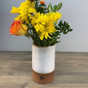 May include: White ceramic vase with a brown base, featuring the number "9" and a heart. The vase is filled with yellow and orange flowers.