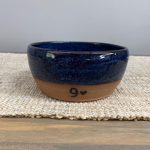 May include: A handmade ceramic bowl with a dark blue glazed interior and a natural clay exterior. The number "9" and a heart symbol are painted on the exterior. The bowl is on a woven mat.