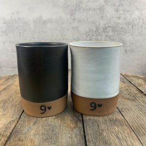 May include: Two ceramic mugs with a brown rim and a black heart symbol. The mug on the left is black with a brown rim and the mug on the right is white with a brown rim. Both mugs have the number "9" written on the front.