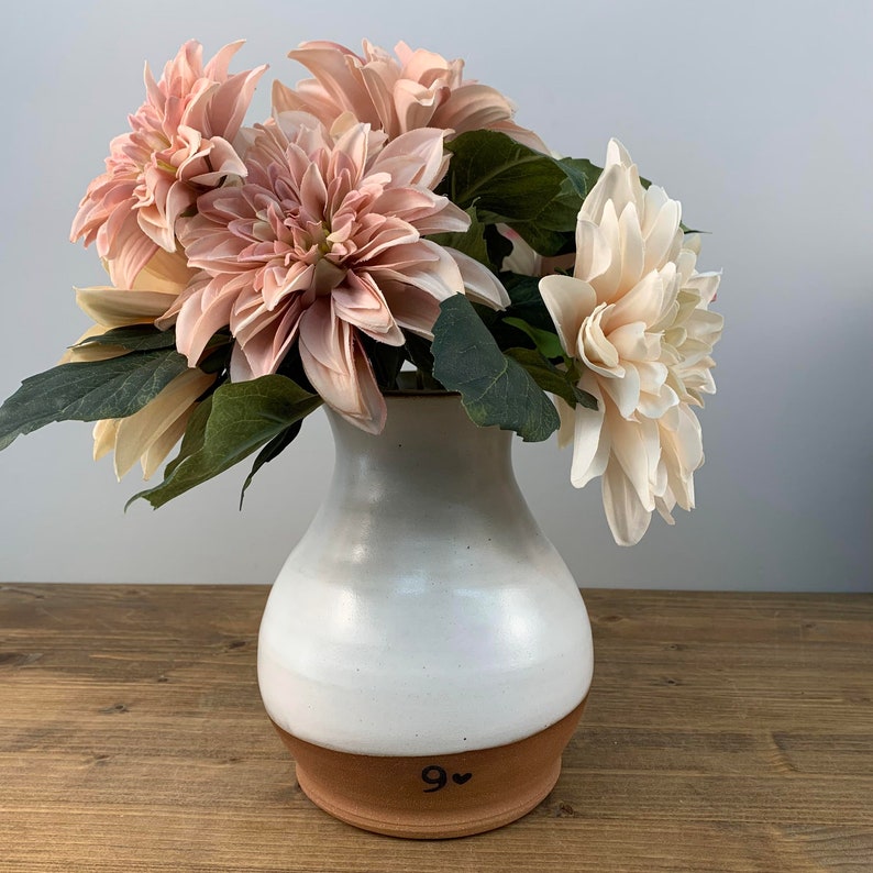 May include: A white ceramic vase with a brown rim and the number "9" with a heart next to it. The vase is filled with a bouquet of pink and cream colored dahlias.