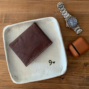 May include: A brown leather wallet with a white tray and a silver watch on a wooden surface. The tray has the number 9 and a heart drawn on it.
