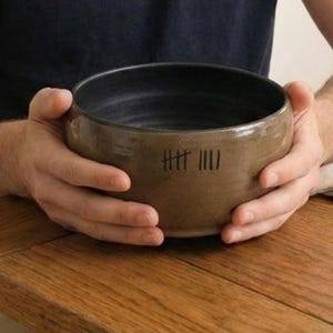 May include: A handmade ceramic bowl with a dark interior and a tan exterior. The bowl is held by two hands and has a series of tally marks etched on the side. The bowl is sitting on a wooden table.