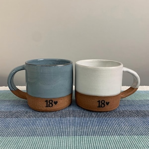 May include: Two ceramic mugs with handles, one in dusty blue and one in off-white, both featuring a raw terracotta base. Each mug is stamped with the number 18 followed by a small heart symbol in black ink on the lower terracotta section.