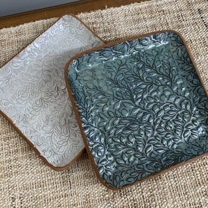 May include: Two square ceramic dishes with rounded corners and a raised leaf pattern. One dish is finished in a glossy white glaze, while the other features a deep blue-green glaze. Both items have exposed brown clay edges along the rims.