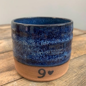 May include: A handmade ceramic mug with a dark blue, textured glaze on the upper portion and a natural clay base. The number "9" and a heart are painted in black on the base. The mug is sitting on a wooden surface.