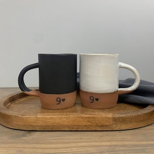 May include: Two ceramic mugs, one black and one white, with a terracotta-colored base. Each mug has a curved handle and the number "9" with a heart symbol on the base. The mugs are on a wooden tray.
