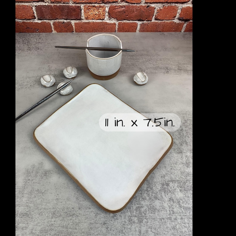 May include: A white ceramic plate with a brown rim, measuring 11 inches by 7.5 inches, is displayed alongside a small ceramic cup, chopsticks, and several small, round ceramic pieces. The items are arranged on a gray surface.
